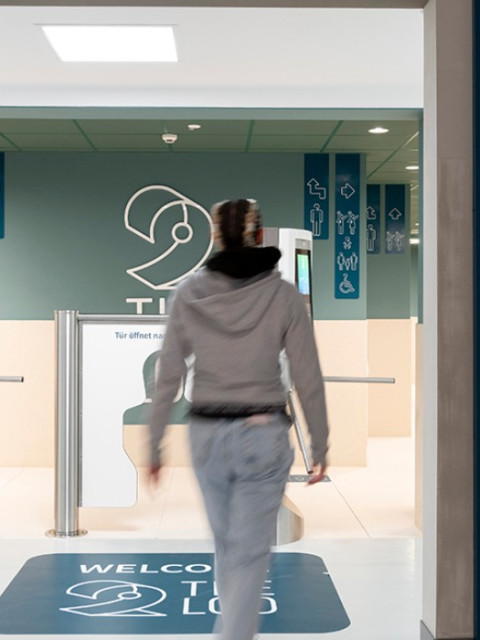 A person walks towards a restroom entrance, with directional signage on the walls indicating bathroom options and accessibility features.