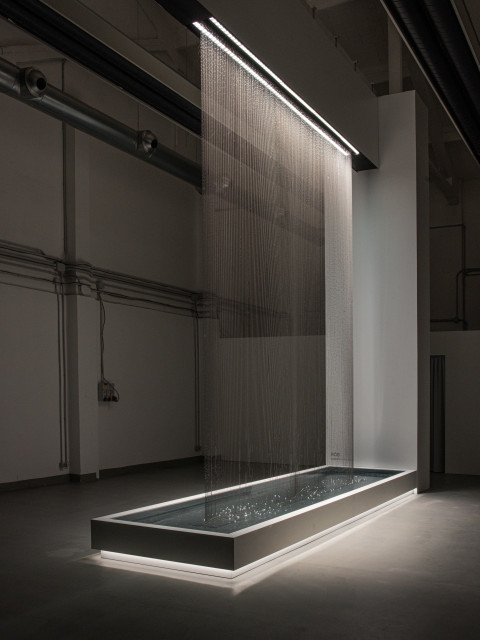 A contemporary art installation featuring a rectangular basin with water illuminated by lights, under a vertical sheet of cascading water.
