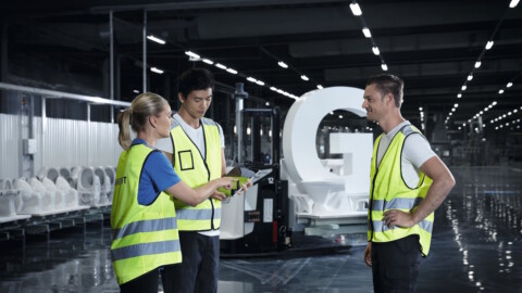 Drei Techniker in Warnwesten diskutieren die Baupläne in einer modernen Fertigungsanlage vor weissen Keramiken.