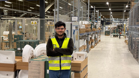 A person wearing a yellow reflective vest stands with arms crossed in a warehouse aisle lined with boxes and equipment.