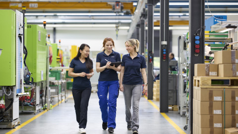 Three women in work uniforms walking together in a factory, discussing and holding a tablet.