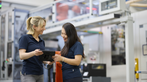 Two women in dark polo shirts collaboratively reviewing information on a tablet in an industrial setting with machinery in the background.