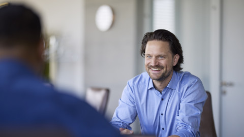 A man in a light blue shirt smiles while engaged in a conversation at a conference table, with another person partially visible.