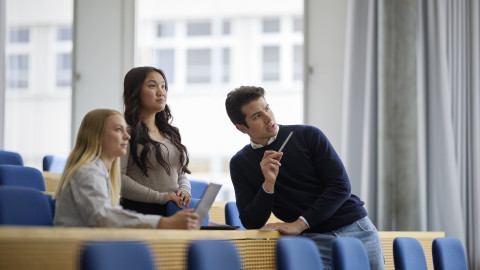 Three young adults in a modern classroom setting, engaged in discussion with notebooks and pens.