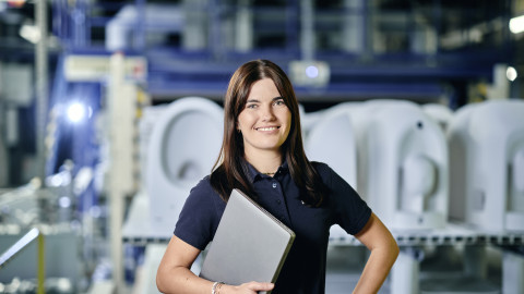 A woman in a polo shirt holds a laptop while standing in an industrial setting.