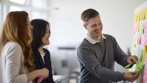Three professionals engaging in a collaborative brainstorming session in an office, with one person attaching notes to a board.