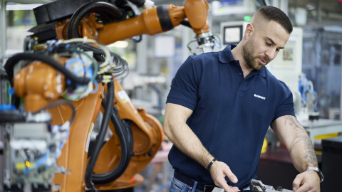 A man in a blue polo shirt works with machinery while a robotic arm is positioned nearby in a manufacturing setting.