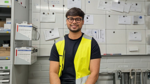 A person wearing a yellow safety vest and glasses, sitting in front of a wall with documents and electrical panels.