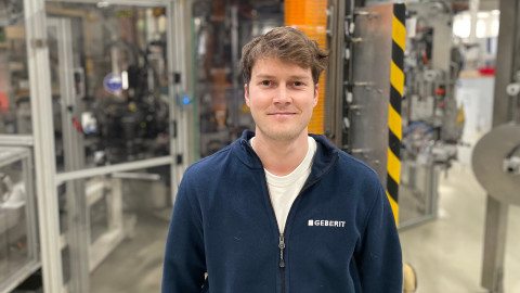 A young man standing in an industrial setting with machinery in the background, wearing a dark blue jacket.