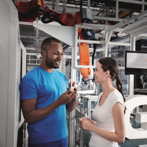 A man and a woman are conversing in a production facility. A white ‘G’ is visible in the background.