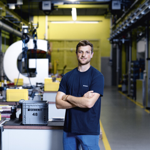 A young man in a dark blue shirt and jeans with crossed arms in a brightly lit factory.