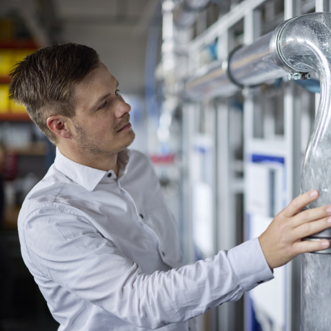 Ein Mann in einem weissen Hemd betrachtet und berührt eine grosse, transparente Rohrleitung in einem technischen Umfeld.