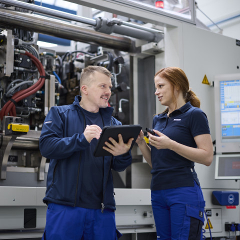 Zwei Techniker in Arbeitskleidung diskutieren vor einer grossen Maschinenanlage, während einer ein Tablet hält.