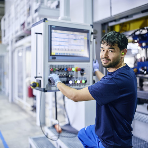 Ein Mann bedient eine Maschine in einer modernen Produktionshalle, mit einem Bedienfeld voller Knöpfe und einem Bildschirm.