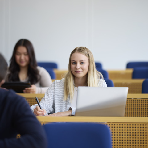 Eine Gruppe von Studierenden in einem modernen Klassenraum, eine blonde Frau lächelt und arbeitet an einem Laptop.