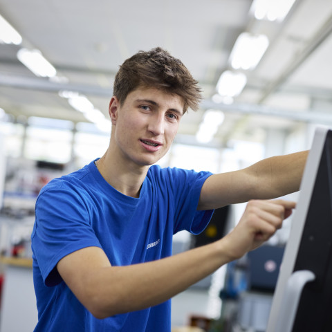 Junger Mann in blauem T-Shirt bedient einen Bildschirm in einer modernen Werkstatt.