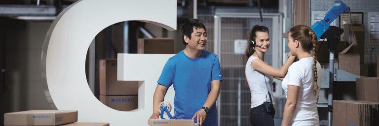 Three employees are working in a storage room filled with cardboard boxes and a large stylised letter ‘G’.