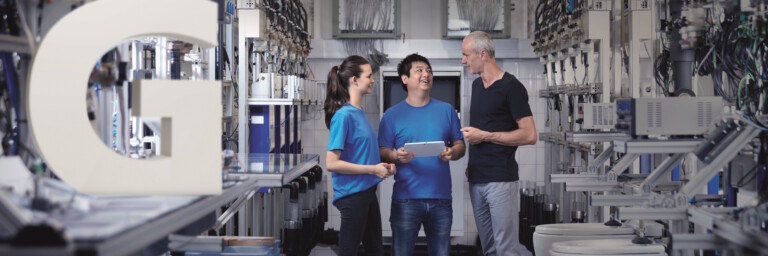 Three technicians are in a sanitary testing facility. A white letter ‘G’ is placed in the left-hand section of the image.