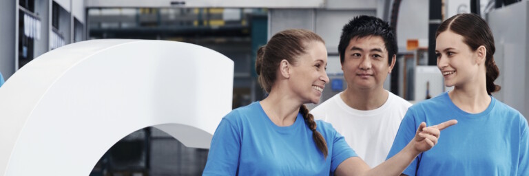 Three smiling employees in blue shirts stand next to a large white G sculpture.