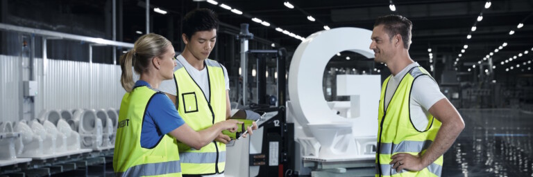 Drei Techniker in Warnwesten diskutieren die Baupläne in einer modernen Fertigungsanlage vor weissen Keramiken.