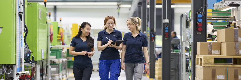 Drei Frauen in Arbeitskleidung gehen lachend in einer Fabrikhalle, umgeben von Maschinen und Kartons.
