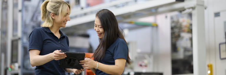 Zwei Frauen in Arbeitskleidung lächeln und schauen auf ein Tablet in einer modernen Fertigungsumgebung.
