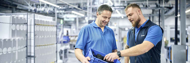 Zwei Männer in blauen Shirts stehen in einer Lagerhalle und einer hält einen blauen Behälter.