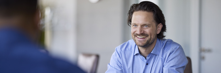 Ein lächelnder Mann im blauen Hemd sitzt an einem Tisch und führt ein Gespräch mit einer anderen Person.