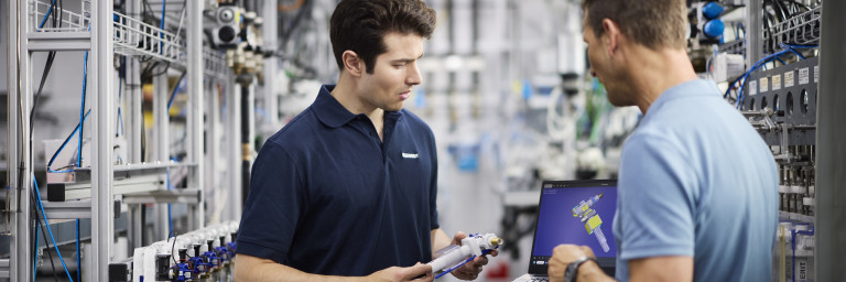 Zwei Männer in einer industriellen Umgebung, einer mit Laptop und der andere mit einem Produkt in der Hand, diskutieren.