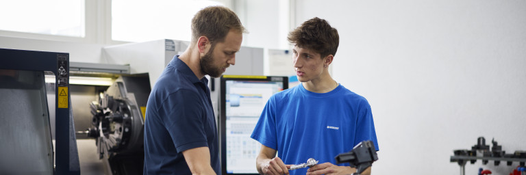 Zwei Personen in einem technischen Umfeld unterhalten sich über Werkstücke an einem Arbeitstisch neben einer Maschinenbedienung.