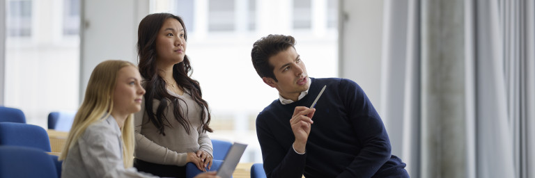 Three young adults in a modern classroom setting, engaged in discussion with notebooks and pens.