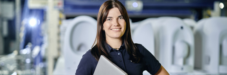 A woman in a polo shirt holds a laptop while standing in an industrial setting.