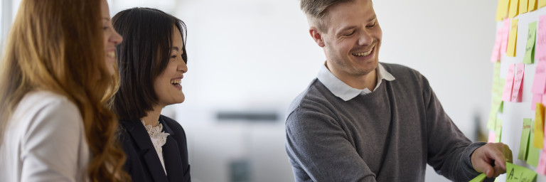 Three professionals engaging in a collaborative brainstorming session in an office, with one person attaching notes to a board.
