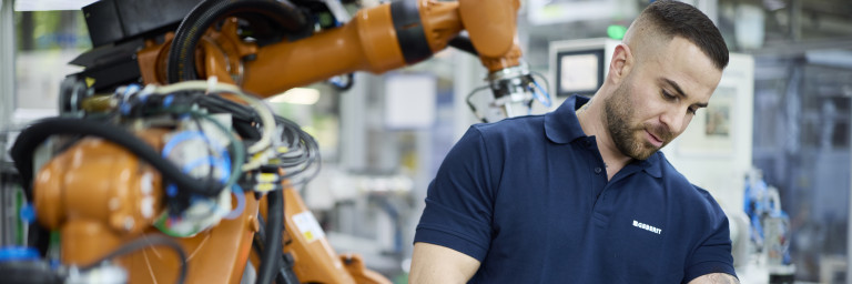 A man in a blue polo shirt works with machinery while a robotic arm is positioned nearby in a manufacturing setting.