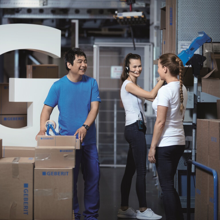 Three employees are working in a storage room filled with cardboard boxes and a large stylised letter ‘G’.