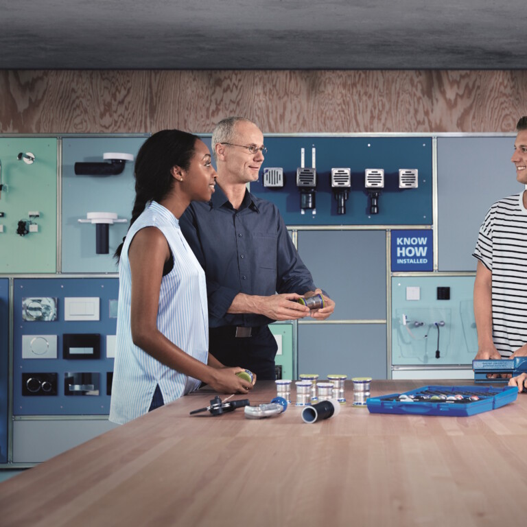 4 Personen stehen um einen Tisch mit Geberit Mapress Produkten und tauschen sich aus. Auf der rechten Seite im Hintergrund steht ein weisses G.