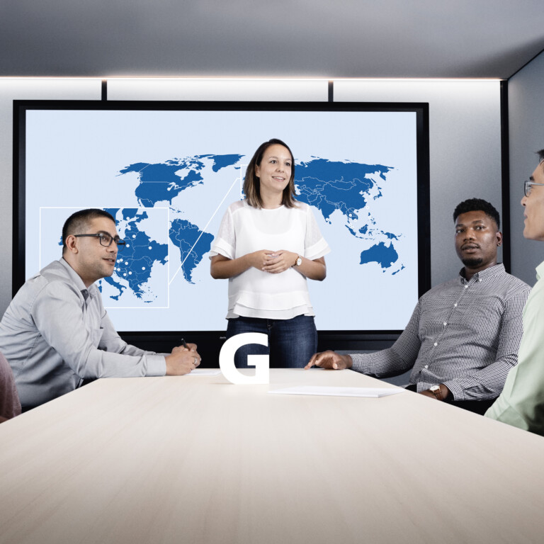 Eine engagierte Frau, die vor einer Weltkarte und vier Zuschauern in einem Besprechungsraum steht.