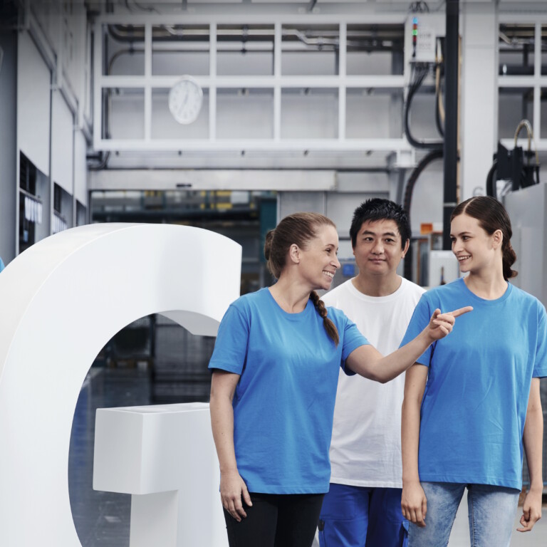 Three smiling employees in blue shirts stand next to a large white G sculpture.