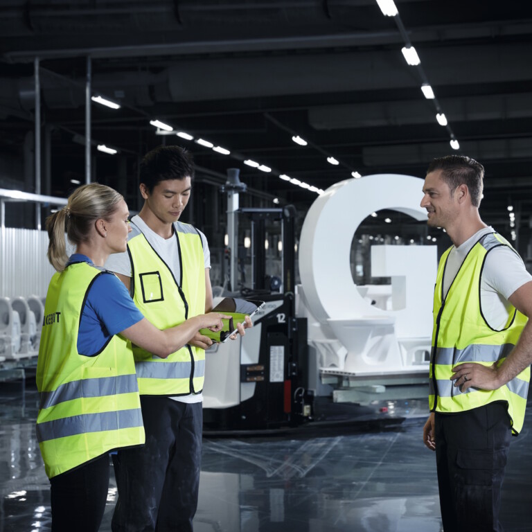 Drei Techniker in Warnwesten diskutieren die Baupläne in einer modernen Fertigungsanlage vor weissen Keramiken.