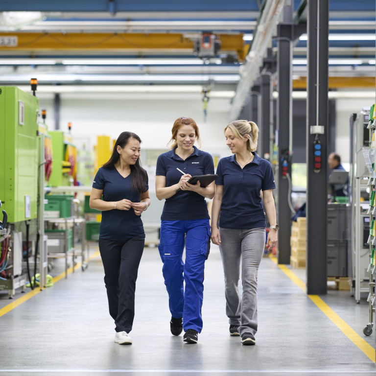 Drei Frauen in Arbeitskleidung gehen lachend in einer Fabrikhalle, umgeben von Maschinen und Kartons.