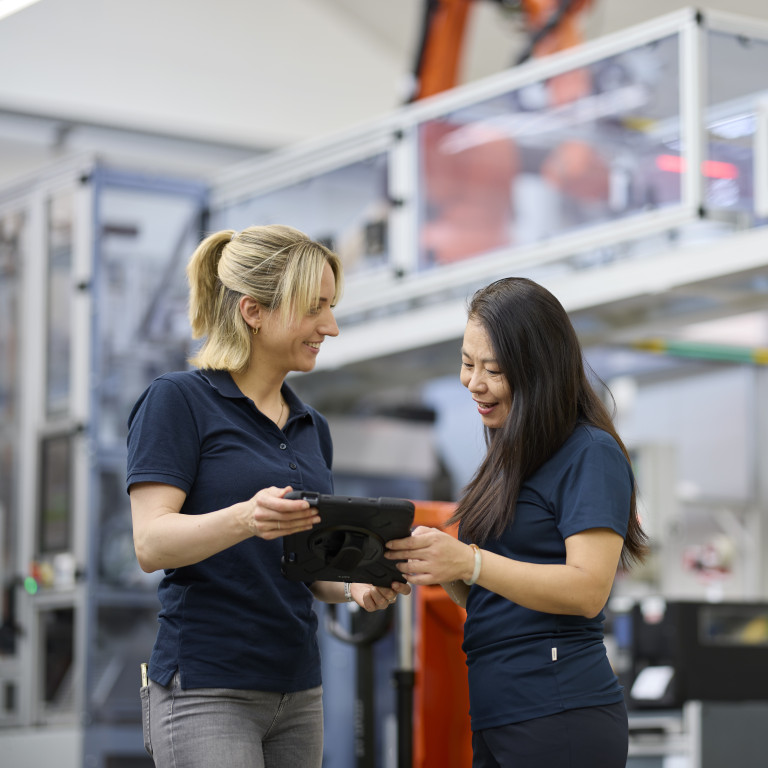Zwei Frauen in Arbeitskleidung lächeln und schauen auf ein Tablet in einer modernen Fertigungsumgebung.