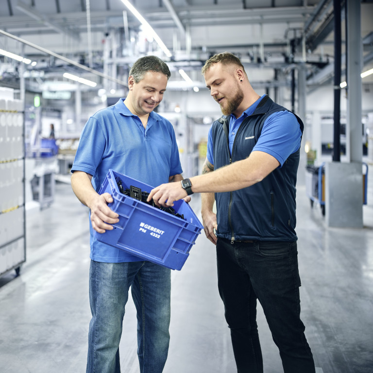 Zwei Männer in blauen Shirts stehen in einer Lagerhalle und einer hält einen blauen Behälter.