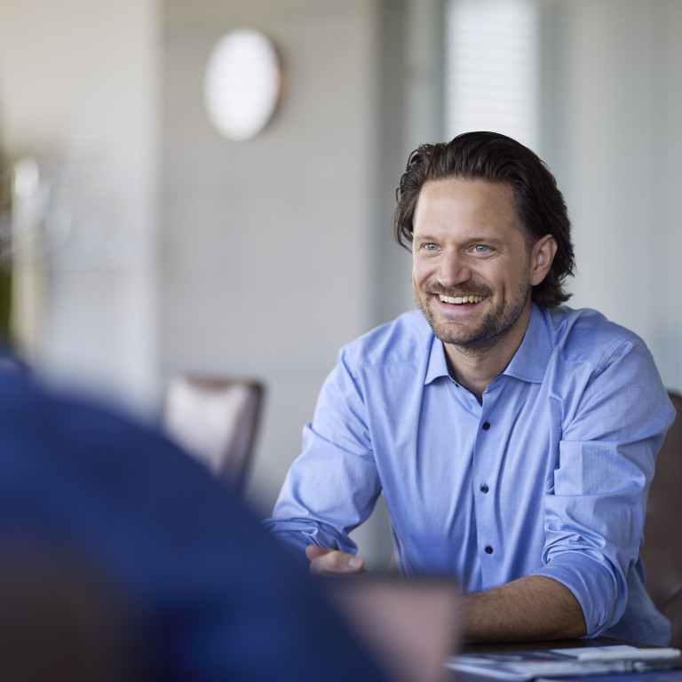 Ein lächelnder Mann im blauen Hemd sitzt an einem Tisch und führt ein Gespräch mit einer anderen Person.
