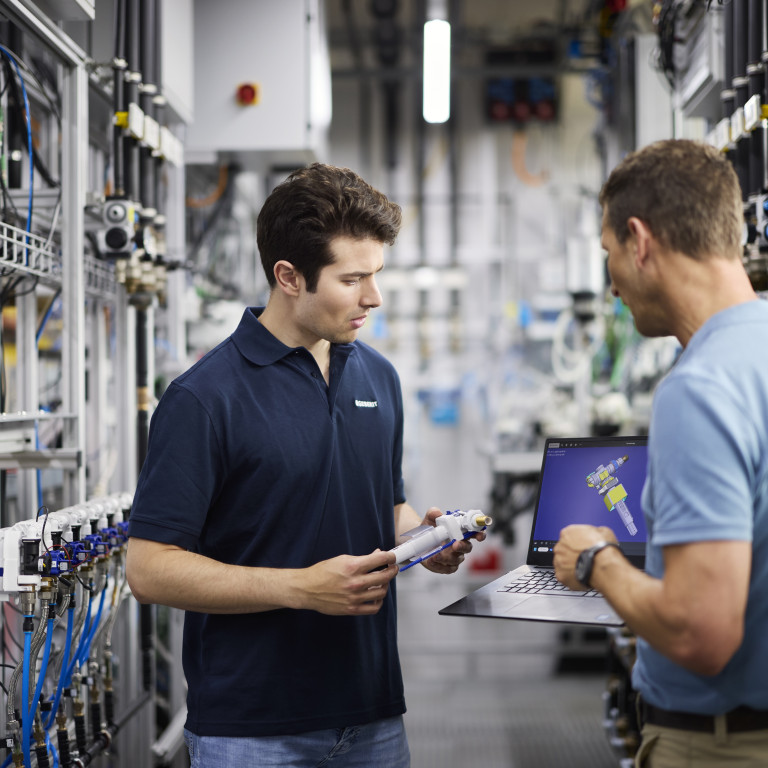 Zwei Männer in einer industriellen Umgebung, einer mit Laptop und der andere mit einem Produkt in der Hand, diskutieren.
