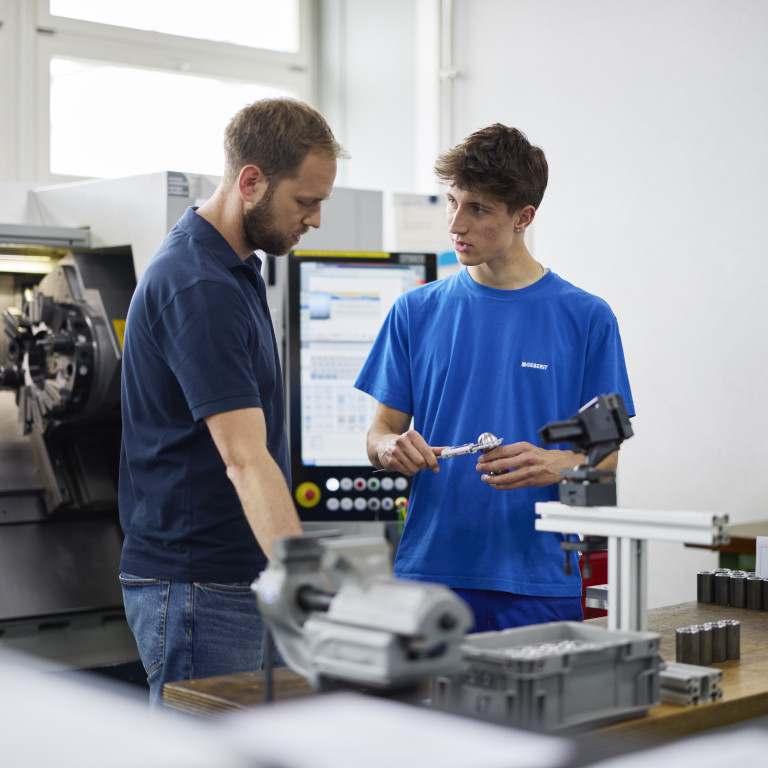 Zwei Personen in einem technischen Umfeld unterhalten sich über Werkstücke an einem Arbeitstisch neben einer Maschinenbedienung.