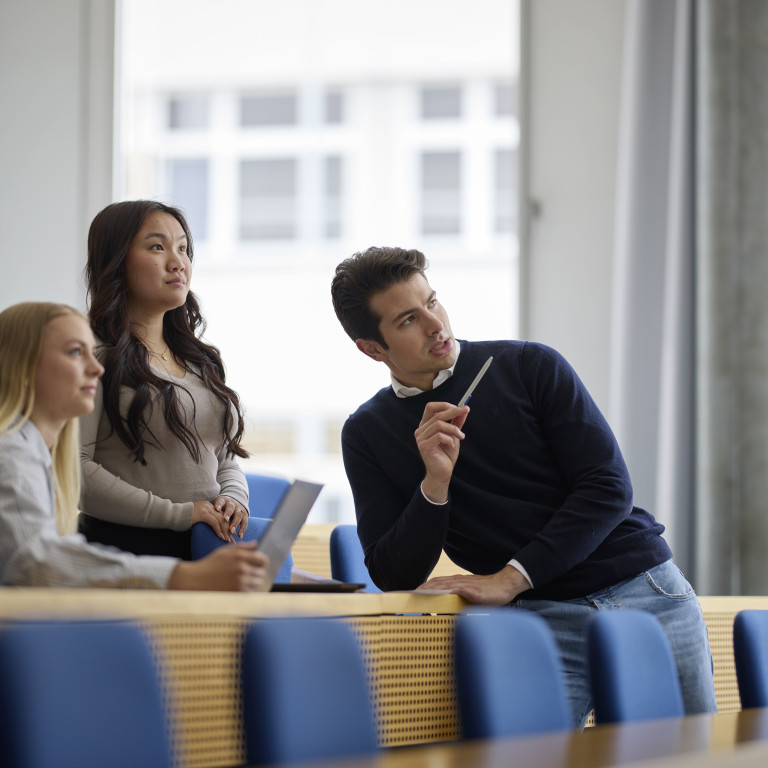 Three young adults in a modern classroom setting, engaged in discussion with notebooks and pens.