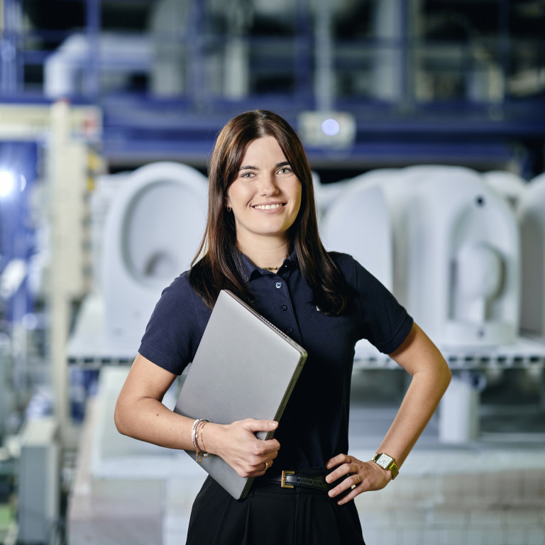 A woman in a polo shirt holds a laptop while standing in an industrial setting.