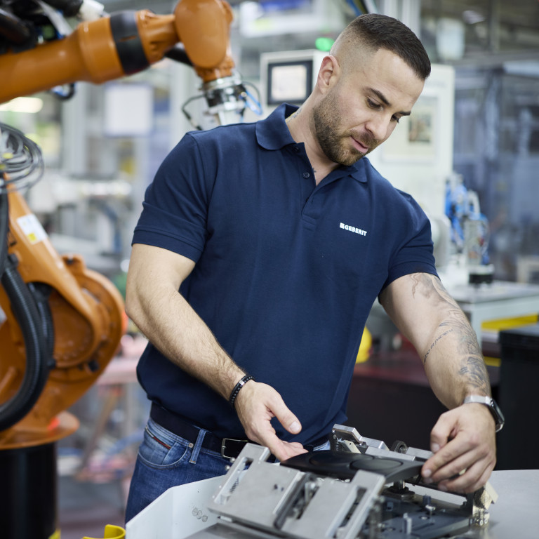 A man in a blue polo shirt works with machinery while a robotic arm is positioned nearby in a manufacturing setting.