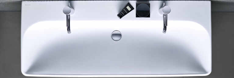 Von oben gesehenes weisses, rechteckiges Waschbecken mit zwei Wasserhähnen und Pflegeprodukten.