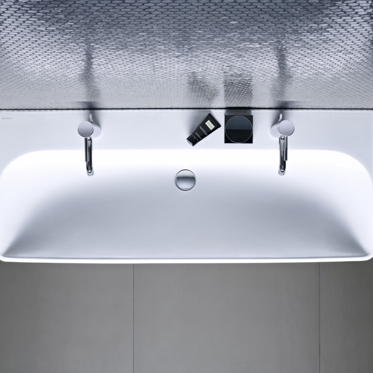 Von oben gesehenes weisses, rechteckiges Waschbecken mit zwei Wasserhähnen und Pflegeprodukten.